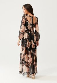 Žena v dlouhých černých květinových šatech s průhlednými dlouhými rukávy, na otevřeném zadním díle svázanými na krku, a v béžových vysokých podpatcích odchází.