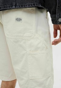 Shorts cargo beige clair en coton avec plusieurs poches et une étiquette tissée. Le tissu est lisse avec un léger éclat.