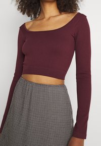 Burgundy ribbed pitkähihaine crop-top, jossa on neliönmuotoinen kaula-aukko, yhdistettynä musta-valkoiseen ruutukuosisiseen hameeseen.
