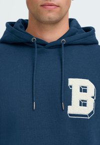 Jeune personne portant un sweat à capuche bleu foncé avec une lettre "B" texturée en blanc sur la poitrine, visible des épaules à la bouche.