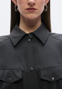 Σκούρο πουκάμισο με κουμπιά, διακοσμημένο με σκληρή γιακά, ασημένια κουμπιά με κλιπ και δύο μπροστινές τσέπες. Λείο ύφασμα με ματ υφή.