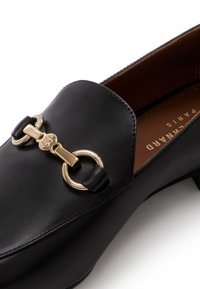 Sorte læder loafers med en guldfarvet hardware accent med cirkulære ringe og en lille kæde detalje. Glat tekstur og lav hældesign.