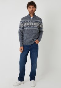 Gebreide pullover in marineblauw met een hoge kraag, met een geometrisch wit en blauw patroon over de borst. Gecombineerd met blauwe jeans en witte sneakers.