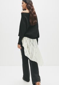 Zwarte oversized trui met geribbelde manchetten, laag over een witte asymmetrische kanten rok. Zwarte flared broeken maken de outfit compleet.