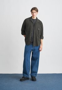 Camisa de flanela preta e verde xadrez sobre uma t-shirt escura, combinada com jeans azuis folgados e calçado preto, em um fundo neutro.