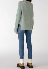 Personne portant un pull en maille vert clair, un jean bleu, des chaussettes crème et des chaussures à plateforme beige, se tenant de dos sur un fond blanc.