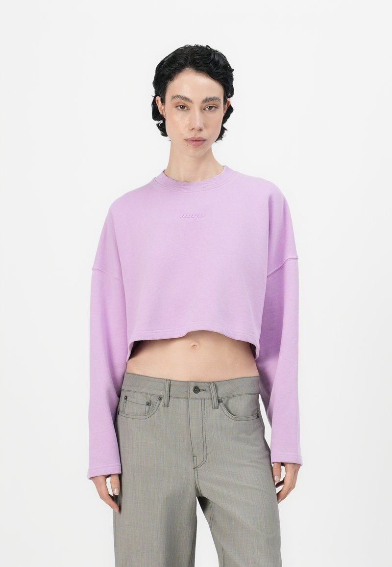 Lila lyhyt hiha sweatshirt, jossa on pitkät hihat ja ribbitetty kaulus. Sisältää hienovaraisen kohokuvioidun logon rinnassa. Yhdistetty harmaisiin housuihin.