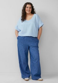 Helles blaues Leinenoberteil mit V-Ausschnitt und aufgerollten Ärmeln, kombiniert mit lockeren, blauen strukturierten Hosen. Das Outfit wird durch lässige Schuhe ergänzt.