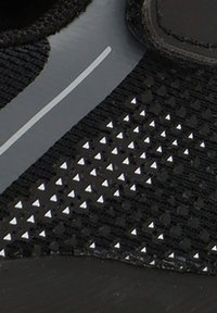 Zapato deportivo negro con una parte superior texturizada que presenta pequeños acentos triangulares blancos y una franja gris suave a lo largo del costado.