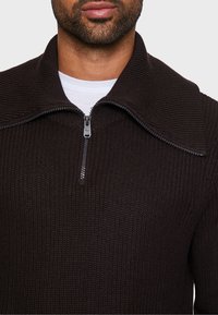 Brauner Strickpullover mit hohem Kragen, ausgestattet mit einem silbernen Reißverschluss am Hals. Die Textur ist gerippt und wirkt warm und strukturiert.