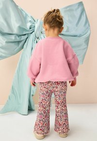 Hanorac roz cu umeri căzuți, asortat cu pantaloni evazați cu flori, în nuanțe de roșu și violet. Fundalul include o mare fundă din material turcoaz.