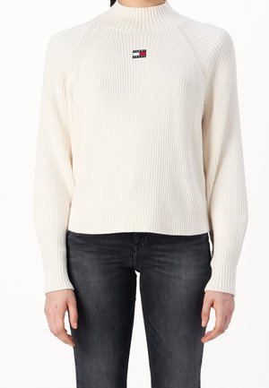 Kobieta ubrana w kremowy prążkowany sweter z logo Tommy Jeans na piersi i ciemnoszare dżinsy, stojąca na białym tle.