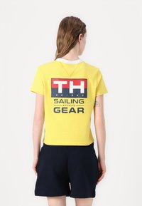 Gul bomulds t-shirt med hvid krave, der har et stort grafisk print på ryggen, som inkluderer marineblå og rød tekst, samt et cropped design.