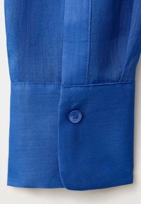 Poignet de chemise en lin bleu avec un seul bouton bleu fermé, montrant les coutures détaillées et la texture du tissu.