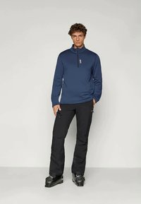 Μαύρο μακρυμάνικο τοπ με φερμουάρ μέχρι το ένα τέταρτο, φορεμένο με μαύρο παντελόνι και μαύρες μπότες σκι; ελαχιστικός σχεδιασμός με μια διακριτική λεπτομέρεια λογότυπου στο στήθος.