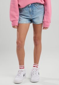 Denim shorts with a faded light blue wash, side pockets, and frayed hem. Paired with white sneakers and red-striped socks.