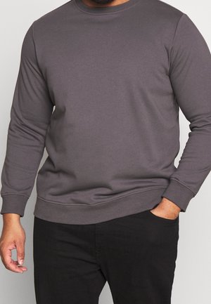 Mann trägt ein schlichtes dunkelgraues Sweatshirt mit langen Ärmeln und Rundhalsausschnitt sowie schwarze Hose, eine Hand in der Tasche, vor neutralem Hintergrund.