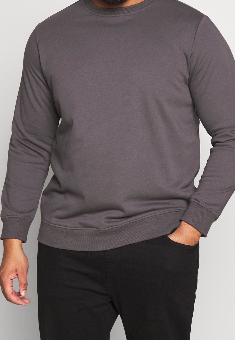 Homme portant un sweat-shirt gris foncé uni à manches longues et col rond, pantalon noir, une main dans la poche, sur un fond neutre.
