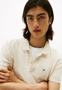 Beige Poloshirt aus strukturiertem Baumwollmaterial, mit klassischem Kragen, drei Knöpfen und einem kleinen gestickten Logo auf der Brust.