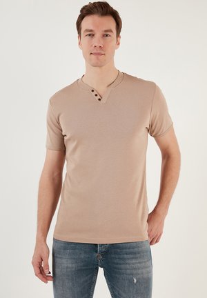 Vaalean beige lyhythihainen T-paita V-kaula-aukolla ja kolmella napilla. Valmistettu pehmeästä kankaasta, jossa on muotoiltu malli ja sileä pinta.