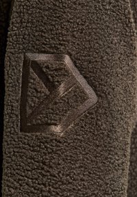 Mørkebrunt fleece-stof med en tekstureret overflade og et geometrisk logo prægning på materialet.
