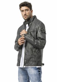 Schwarze Jacke aus Kunstleder mit strukturiertem Kragen, Reißverschlusstaschen und gesteppter Verzierung auf den Schultern; getragen über einem weißen Hemd und blauer Jeans.