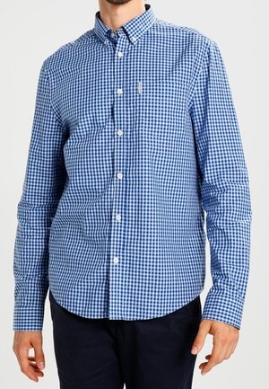 Muž v modro-bílé kostkované košili s dlouhým rukávem, zapínané na knoflíky s límečkem na knoflíky, a černých kalhotách.