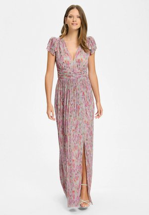 Vrouw die een langroze bloemenjurk draagt met korte pofmouwen, V-hals, geplooide taille en een split aan de voorkant, staand met sandalen.