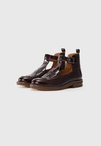 Brown leather shoes with a T-strap design, featuring a woven pattern on the front and a small buckle. Flat, textured rubber sole.