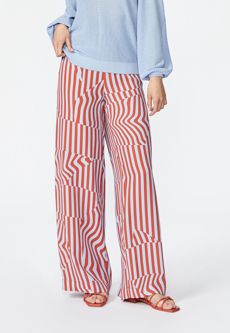 Femme portant un pantalon à jambes larges avec des rayures verticales ondulées rouges et blanches, associées à un pull bleu clair et des sandales rouges à bouts ouverts.