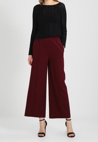 Donna che indossa un maglione nero a maniche lunghe, pantaloni bordeaux a gamba larga e sandali con tacco alto e cinturino alla caviglia neri, in piedi davanti a uno sfondo neutro.