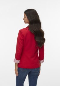 Veste rouge à la coupe ajustée et aux manches trois-quarts, avec des poignets rayés. Portée sur un jean bleu, vue de dos.