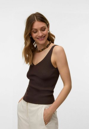 Kobieta z falującymi włosami do ramion, ubrana w brązowy bezrękawnik w prążki, beżowe spodnie, koralikowy naszyjnik i kolczyki koła, uśmiechająca się z zamkniętymi oczami.