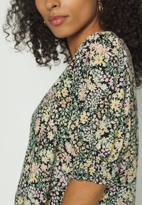 Blusa preta com estampa floral, apresentando mangas curtas em balão com punhos elásticos, um corte solto e um padrão de flores rosas e amarelas sobre um fundo verde.