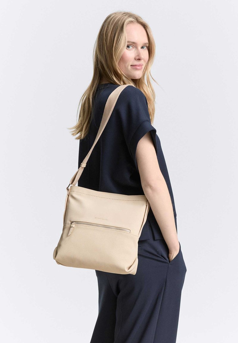 Donna con outfit blu navy che porta una borsa a tracolla beige con tasca frontale con cerniera, che guarda oltre la spalla con la mano nella tasca.
