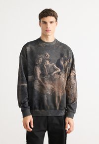 Ung mand iført en mørk sweatshirt med et vintage-inspireret maleri af tre kvinder og en mand i formelt tøj på fronten, stående mod en hvid baggrund.