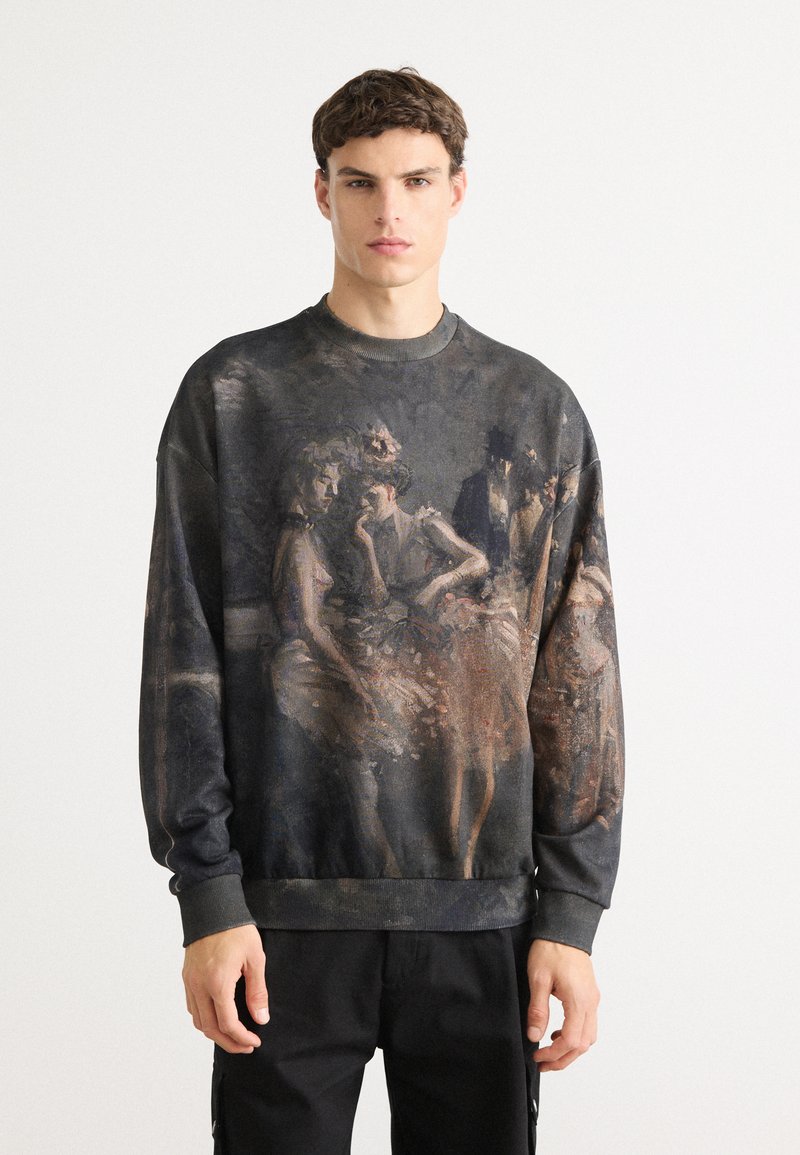 Ung mand iført en mørk sweatshirt med et vintage-inspireret maleri af tre kvinder og en mand i formelt tøj på fronten, stående mod en hvid baggrund.
