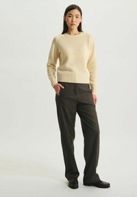Pull en maille beige avec ourlet côtes, associé à un pantalon rayé foncé. L'ensemble présente une coupe décontractée et un design simple. Des chaussures marron complètent le look.
