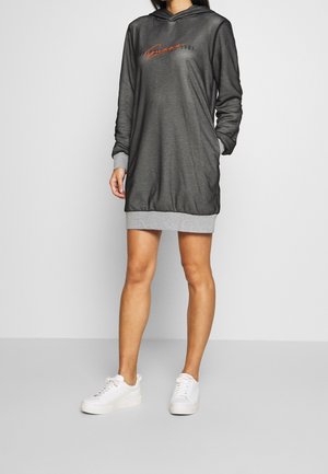 Kobieta ubrana w ciemnoszarą sukienkę typu sweatshirt z kapturem z logo "Guess 1981", szarymi mankietami i dołem oraz białymi trampkami, stojąca.