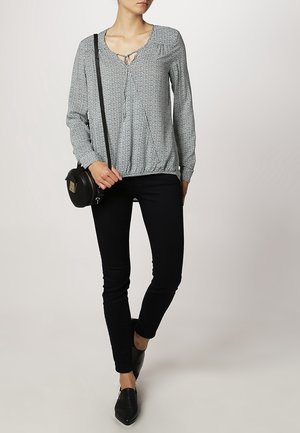 Sort og hvid mønstret bluse med V-udskæring, elastisk kant, og lange ærmer, parret med sorte tætsiddende bukser og flade sorte sko.