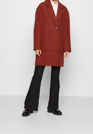 Manteau en laine couleur rouille avec col à revers, fermeture par un seul bouton et deux poches avant, assorti à un pantalon noir évasé et des mocassins brillants.