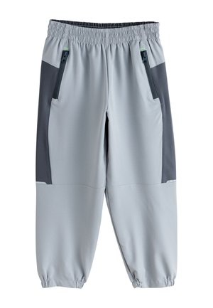 Personne portant un pantalon gris clair avec des poignets élastiques et des baskets blanches Adidas à rayures noires sur un fond neutre.