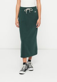 Kvinne i mørkegrønn midi-skirt med snor i livet og lommer, kombinert med svarte høy-snøring sneakers med hvite snørebånd.
