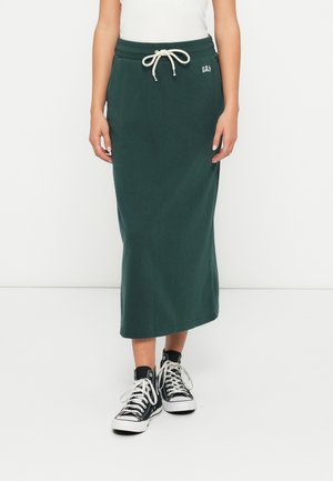 Femme portant une jupe midi vert foncé avec une taille à cordon de serrage et des poches, associée à des baskets montantes noires avec des lacets blancs.
