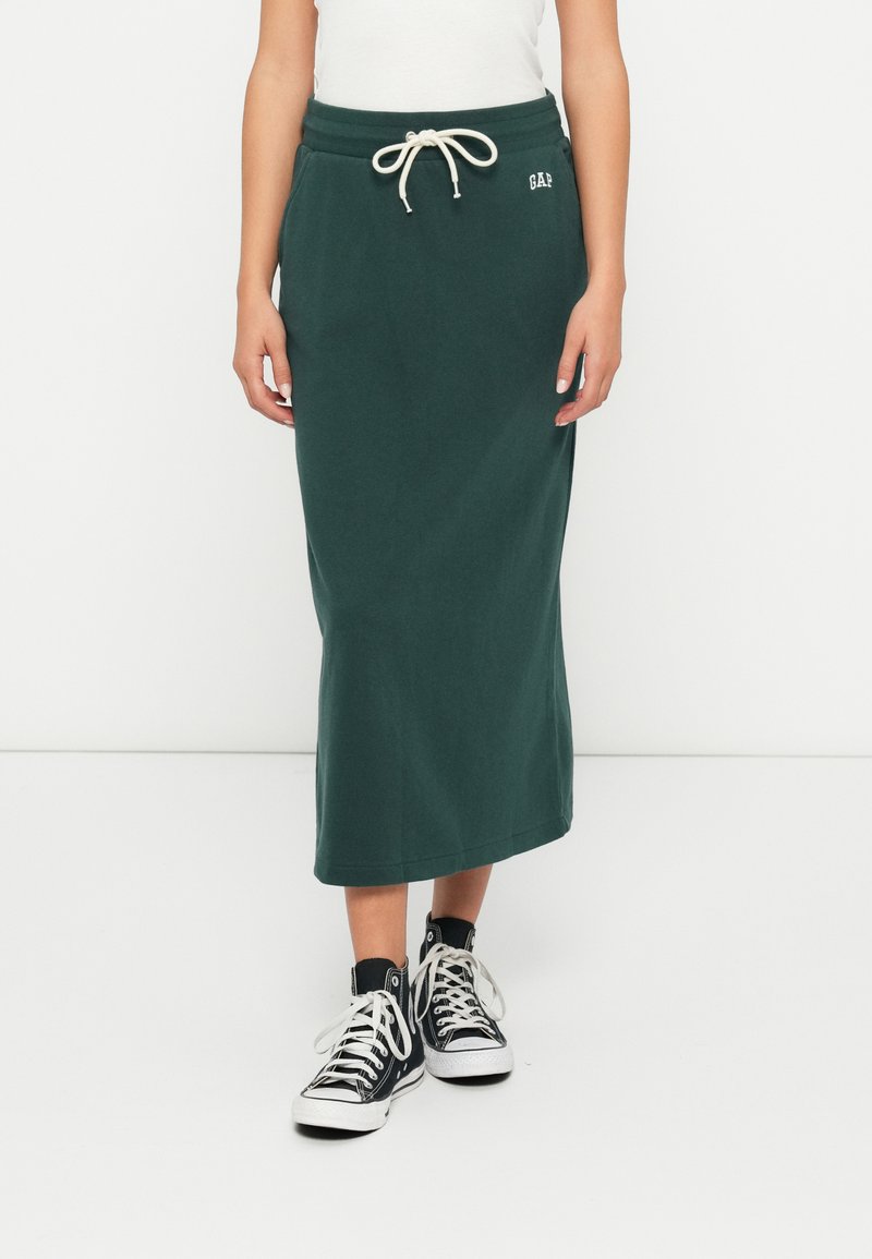 Kvinne i mørkegrønn midi-skirt med snor i livet og lommer, kombinert med svarte høy-snøring sneakers med hvite snørebånd.