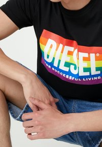 Musta t-paita, jossa on monivärinen sateenkaaren graafinen teksti "DIESEL" ja slogan "KÄYTÄN MENESTYKSEKÄS ELÄMÄ." Yhdistetty farkkushortsien kanssa.