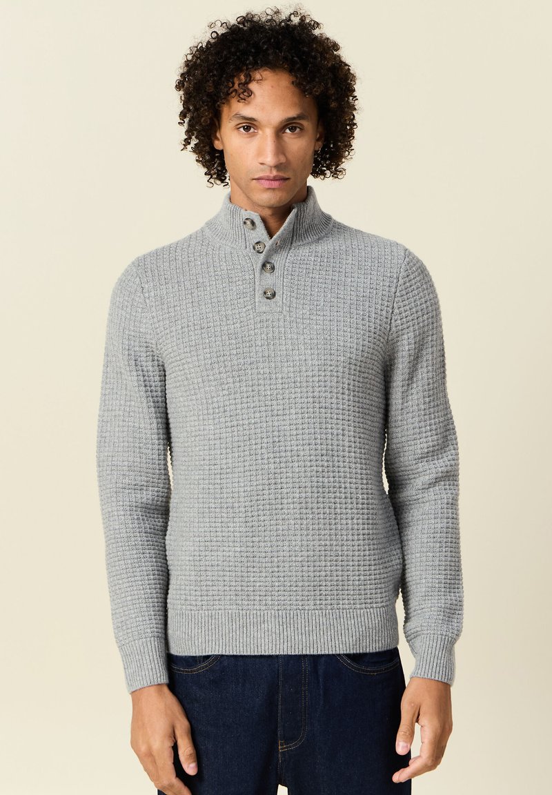 Suéter de punto gris con un acabado texturizado, cuello alto y tapeta de tres botones; puños y dobladillo acanalados, combinado con jeans de mezclilla oscura.