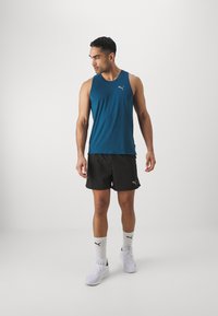 Camiseta atlética sin mangas en color verde azulado con logo, combinada con pantalones cortos negros. Calcetines blancos y zapatillas deportivas completan el conjunto. Tela suave, ajuste casual.