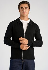 Czarny kardigan na suwak z kołnierzem, ściągaczami przy mankietach i dolnym obrębie, wykonany z teksturowanego materiału; noszony na białej koszuli, o luźnym kroju.