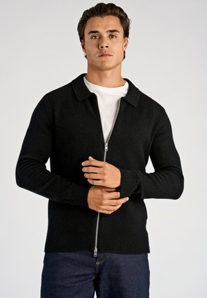 Schwarze Strickjacke mit Reißverschluss, einem Kragen sowie gerippten Bündchen und Saum, gefertigt aus strukturiertem Material; getragen über einem weißen Hemd, mit lässigem Schnitt.